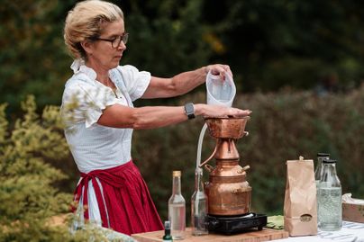 Eine Frau bereitet mit einem Kupfergerät eine Flüssigkeit vor. Sie trägt eine weiße Bluse und einen roten Rock und wirkt konzentriert bei der Arbeit im Freien.