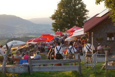 Eine belebte Terrasse mit Gästen, die unter roten Sonnenschirmen sitzen. Im Hintergrund sind Berge und ein malerischer Sonnenuntergang zu sehen.