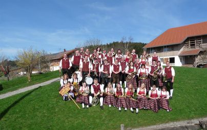 Eine große Gruppe von Musikern steht auf einer Wiese, umgeben von hübschen Gebäuden. Die Mitglieder tragen traditionelle Trachten und halten verschiedene Musikinstrumente in den Händen.