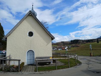 Eine kleine Kirche steht am Wegesrand, umgeben von einer malerischen Landschaft. Der Himmel ist hell und bewölkt, was zu einer ruhigen Atmosphäre beiträgt.