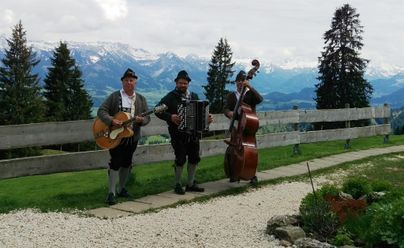 Drei Musiker in traditioneller Tracht spielen in einer malerischen Berglandschaft. Im Hintergrund sind hohe Berge und bewaldete Flächen sichtbar.