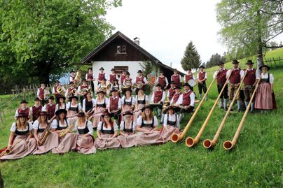 Eine Gruppe von Musikanten in traditioneller Tracht steht vor einem charmanten Fachwerkhaus. Sie halten lange Blasinstrumente und sind von grüner Natur umgeben.