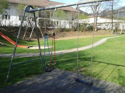 Eine Schaukel auf einem Spielplatz mit Rasen und einem Rutschenbereich im Hintergrund. Die Umgebung ist grün und sonnig.