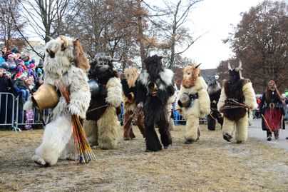 Eine Gruppe von Menschen in traditionellen Kostümen aus Tierfellen marschiert durch eine Straße. Im Hintergrund sind Zuschauer versammelt, die den Umzug beobachten.