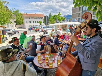 Eine lebhafte Veranstaltung im Freien mit Musikern, die traditionelle Instrumente spielen. An den Tischen sitzen viele Menschen und genießen Getränke.