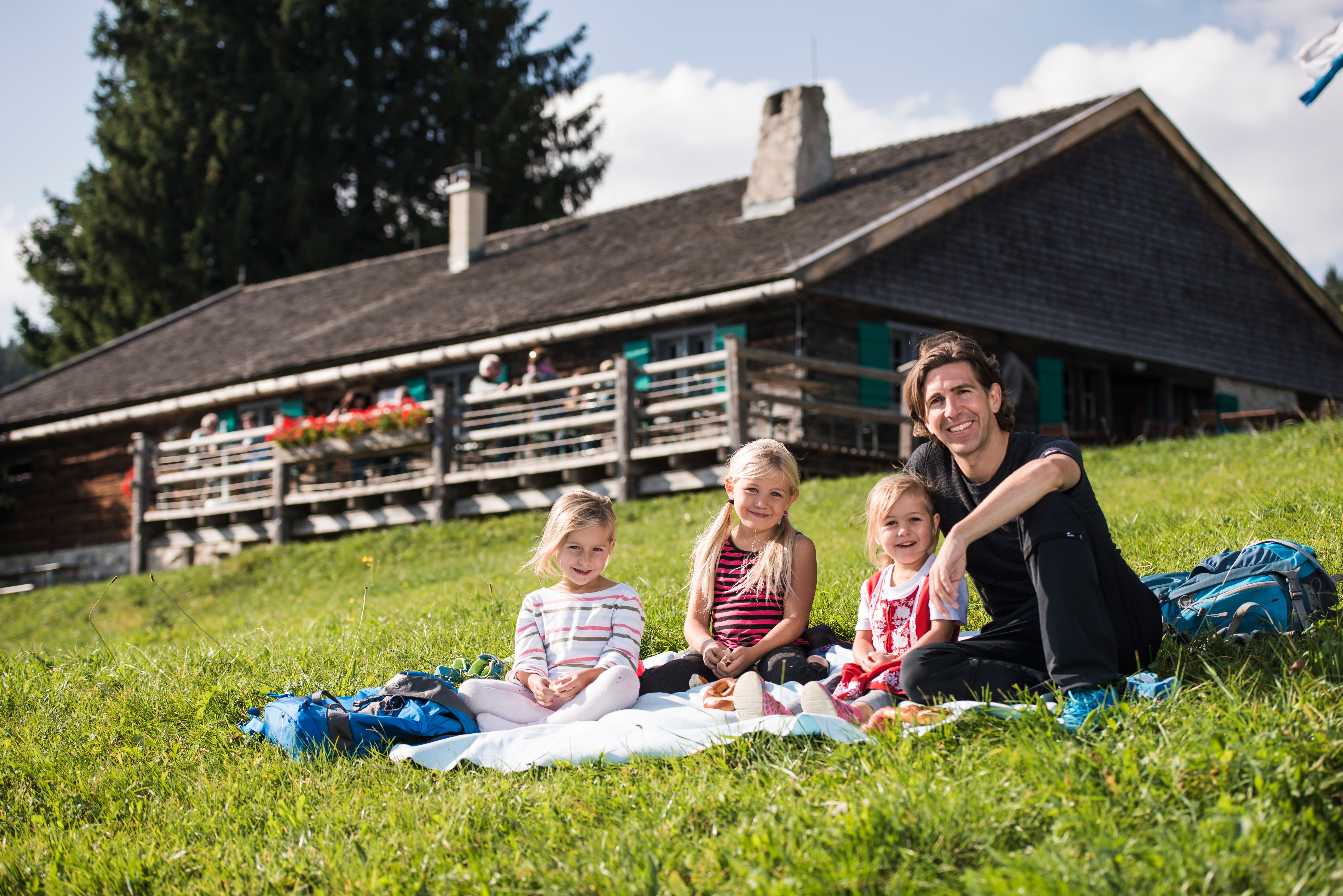 Ein Vater sitz mit seinen drei Mädchen auf einer Picknickdecke vor der Höfle Alpe am Bergbauernmuseum in Diepolz.