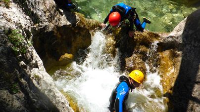 Zwei Personen in Neoprenanzügen klettern über Felsen in einem Wasserfall. Sprudelndes Wasser umgibt sie in einer natürlichen Umgebung.