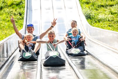 Eine Familie hat Spaß auf einer langen Rutsche in einem Freizeitpark. Sie fahren in zwei Bahnen und lächeln.
