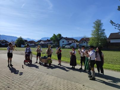 Eine Gruppe von Musikern in Tracht spielt in einer ländlichen Umgebung. Im Hintergrund sind traditionelle Häuser und die Berge zu sehen.