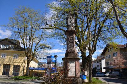 Ein Denkmal steht in einer ruhigen Stadtlandschaft, umgeben von Bäumen und Gebäuden. Der Himmel ist klar und blau.