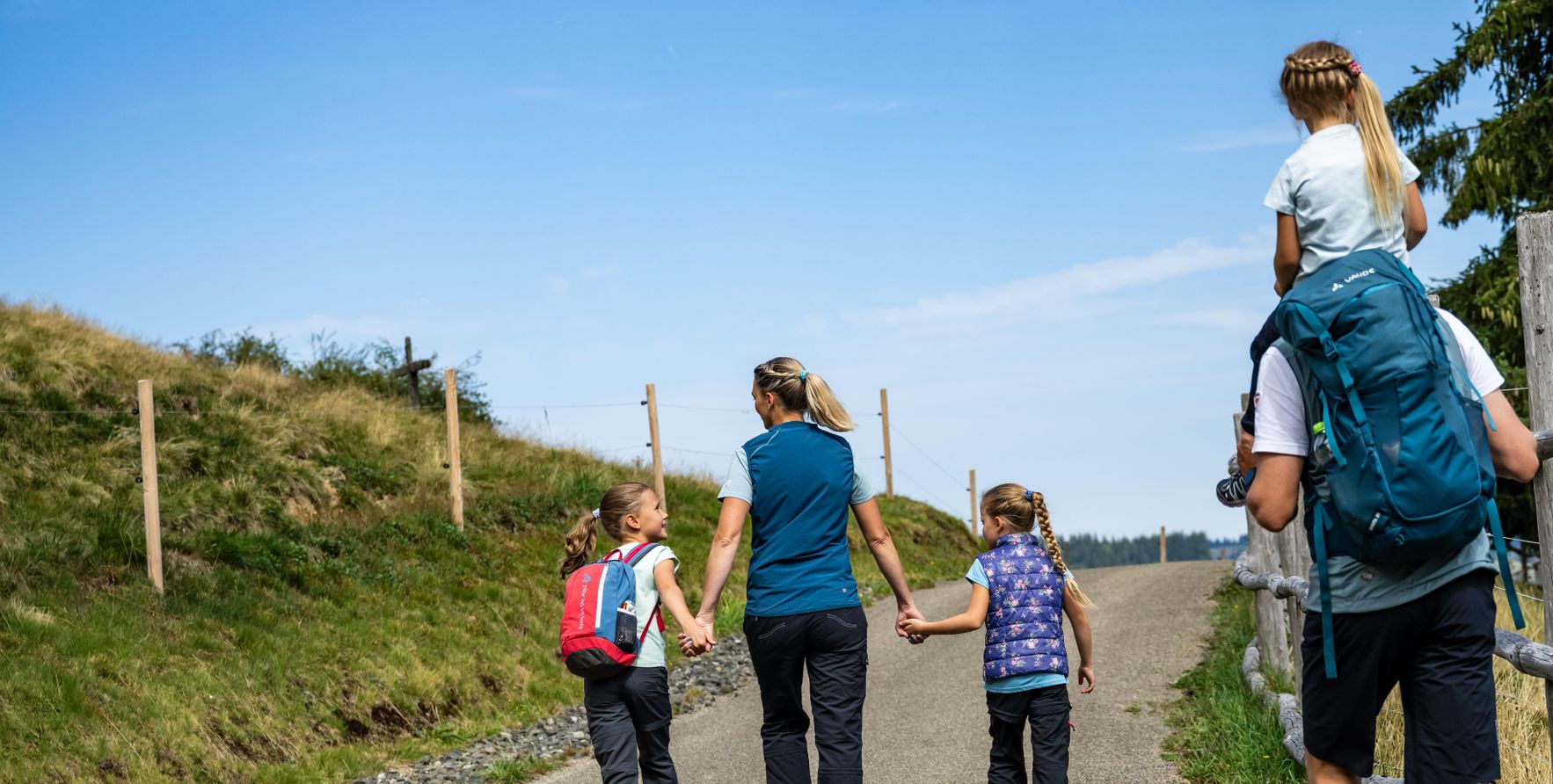 Eine Familie wandert auf einem Weg. Kinder gehen Hand in Hand. Ein Kind sitzt auf dem Rücken eines Erwachsenen. Es ist tagsüber.