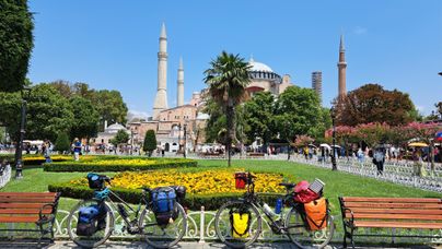 Eine sonnige Szene mit Fahrrädern im Vordergrund, umgeben von bunten Blumen. Im Hintergrund ist die Hagia Sophia und einige Minarette sichtbar.
