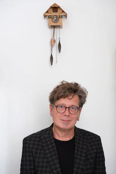 Ein Mann mit Brille und lässiger Kleidung steht vor einer weißen Wand. Im Hintergrund hängt eine traditionelle Kuckucksuhr.