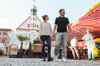 Ein Pärchen spaziert Hand in Hand über einen Markt mit bunten Marktständen. Im Hintergrund ist ein historisches Gebäude zu sehen.