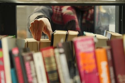 Eine Hand greift nach einem Buch in einer Regalen voller Bücher. Im Hintergrund sind unscharfe Farben eines anderen Buches zu sehen.
