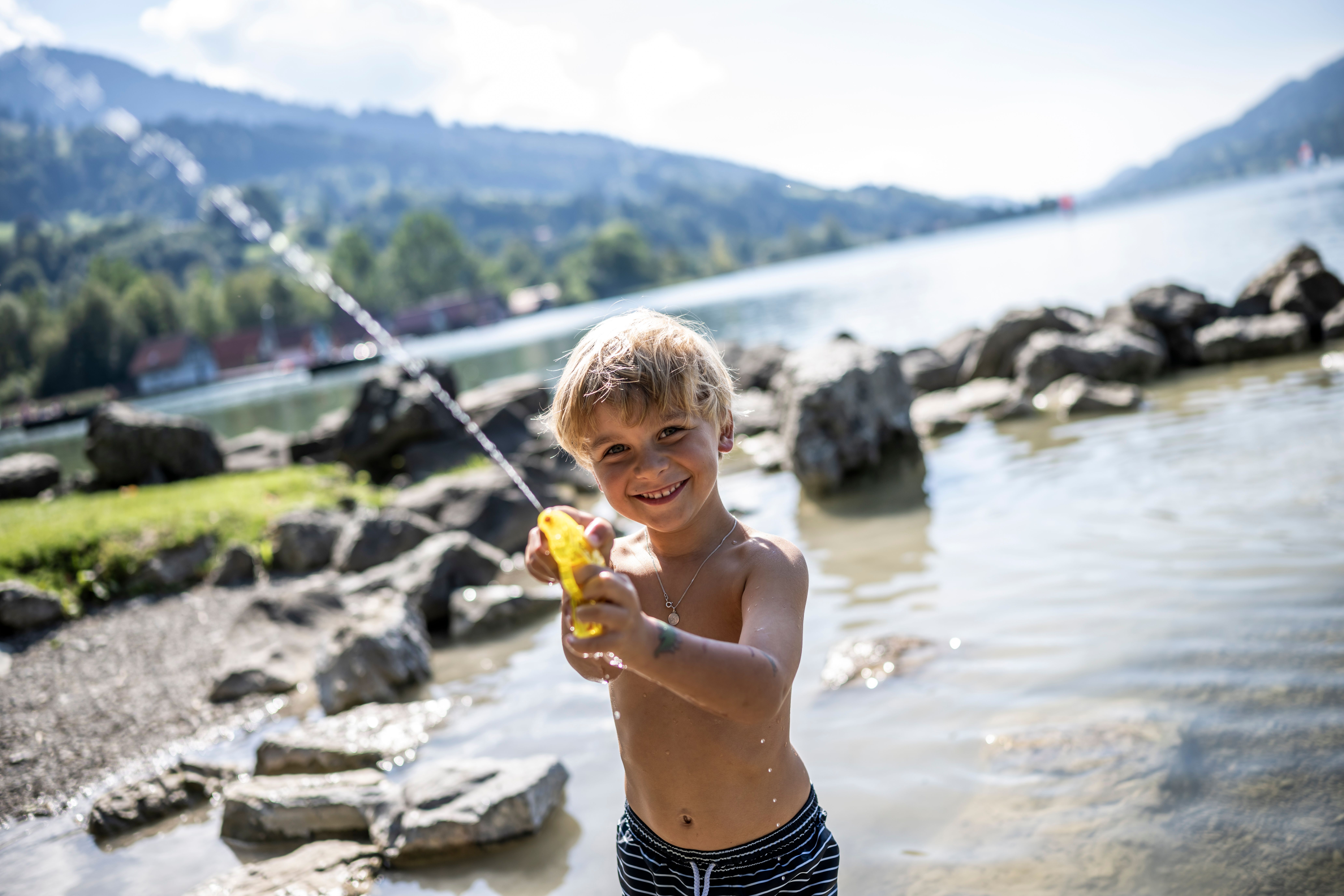 Ein Junge steht im großen Alpsee bei Immenstadt und spielt mit seiner Wasserpistole. Mit einem Umfang von 8 km ist der Natursee umrundet von Grünflächen aber auch Kies, Steinen und Sand.