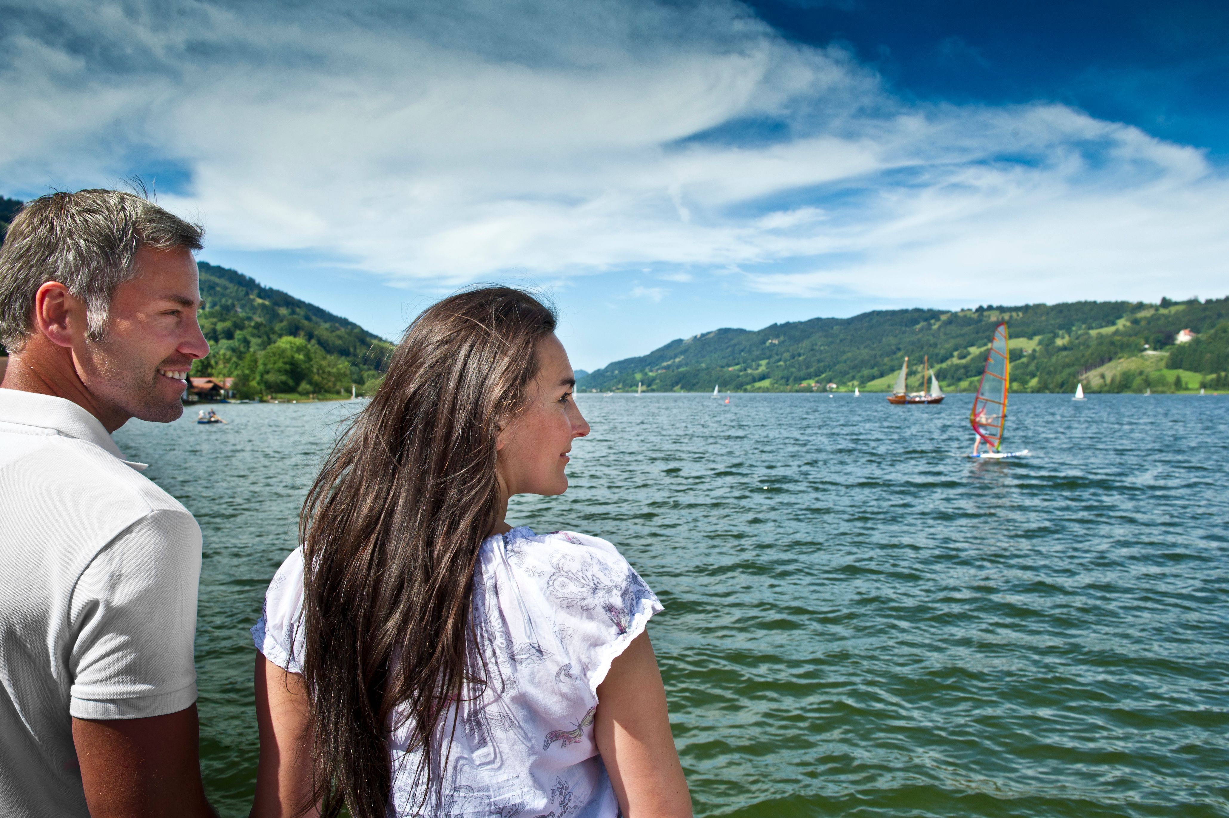 Ein Paar steht am Ufer des Großen Alpsees im Immenstadt und beobachtet Segelboote. Mit einem Umfang von 8 km ist der Natursee umrundet von Grünflächen aber auch Kies, Steinen und Sand.