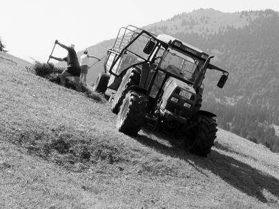 Ein Traktor arbeitet auf einem hügeligen Feld. Im Hintergrund sind Menschen, die Heu sammeln.