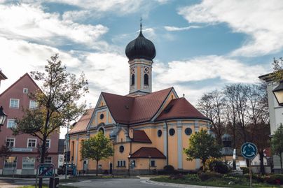 Eine farbenfrohe Kirche mit einer charakteristischen Kuppel steht in einer kleinen Stadt. Umgeben von Bäumen und Wohngebäuden unter einem bewölkten Himmel.