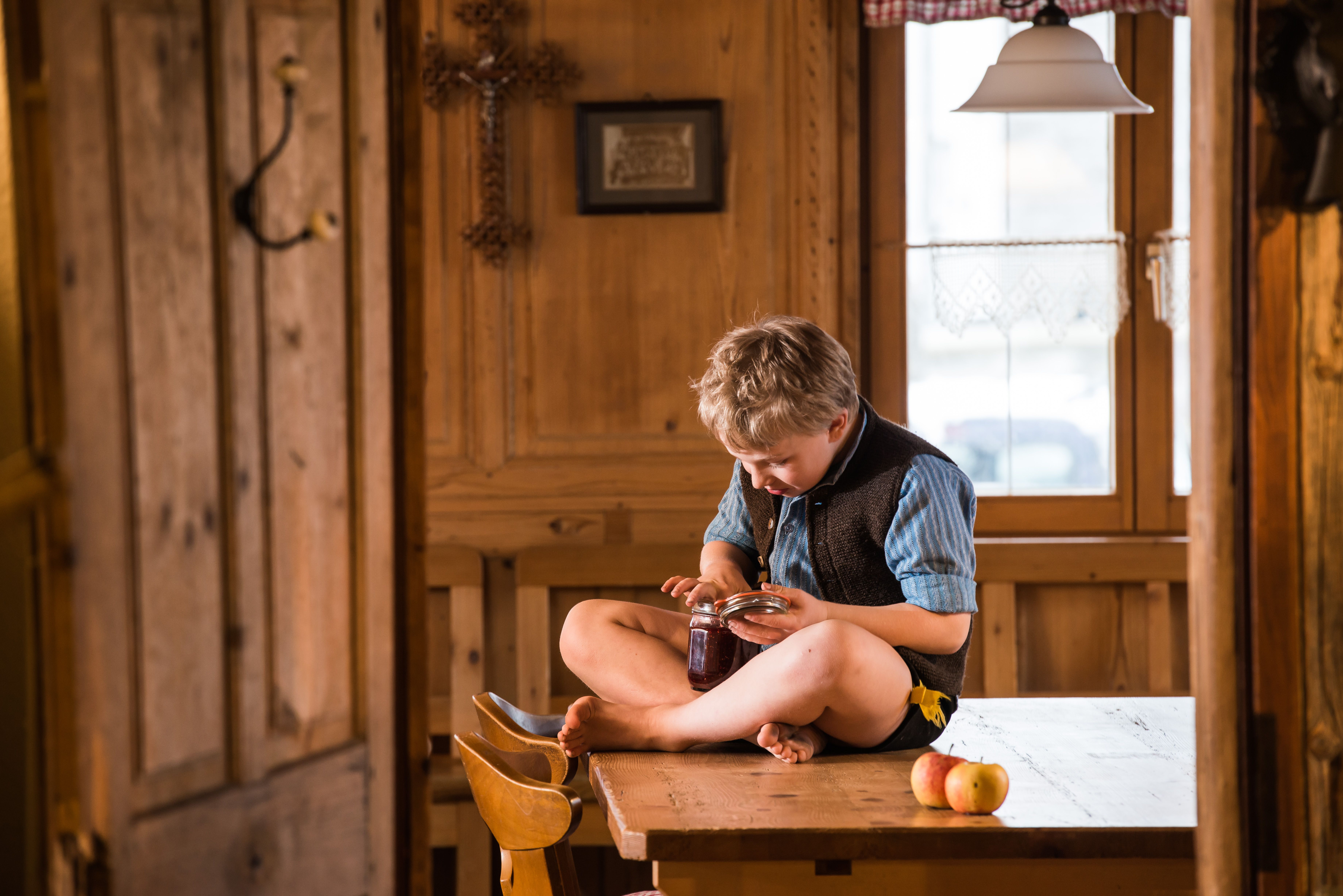 Ein kleiner Junge in Tracht nascht Schokolade mit dem Finger direkt aus dem Glas uns sitzt auf einem Tisch.