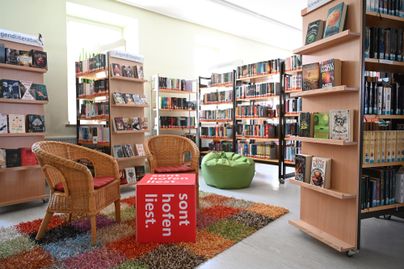 Eine gemütliche Bibliothek mit Regalen voller Bücher. In der Mitte stehen zwei Stühle und ein bunten Teppich.