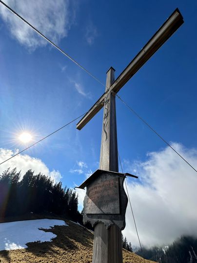 Ein hölzernes Kreuz steht auf einer Bergwiese unter einem klaren, blauen Himmel. Im Hintergrund sind bewaldete Hügel und einige Wolken zu sehen.