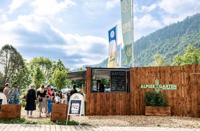 Ein belebter Außenbereich mit einem Holzstand und Menschen, die anstehen. Hintergrund ist eine grüne Landschaft und ein blauer Himmel.