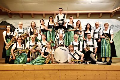 Eine Gruppe von Musikern in Tracht spielt in einer ländlichen Umgebung. Im Hintergrund sind traditionelle Häuser und die Berge zu sehen.