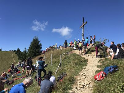 Eine Gruppe von Wanderern versammelt sich um ein Kreuz auf einem Gipfel. Der Himmel ist blau und es gibt viele grüne Wiesen und Bäume in der Umgebung.