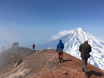 Eine Gruppe von Wanderern befindet sich auf einem Berggipfel mit Blick auf einen schneebedeckten Vulkan. Die Atmosphäre ist klar, aber teilweise neblig, was eine mystische Stimmung erzeugt.